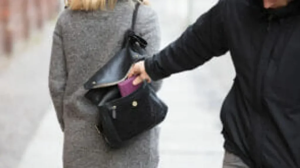 Pessoa vestindo casaco cinza sendo furtada enquanto alguém retira a carteira de sua mochila ou bolsa aberta.