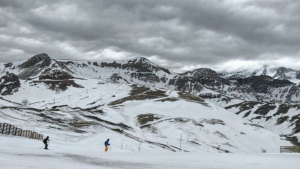 Dois esquiadores em pista com neve escassa e céu carregado, cercados por montanhas rochosas parcialmente cobertas de neve.