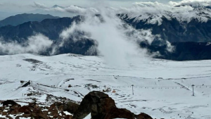Vista ampla de uma estação de esqui coberta de neve com nuvens baixas e montanhas ao fundo, teleféricos visíveis ao longe.