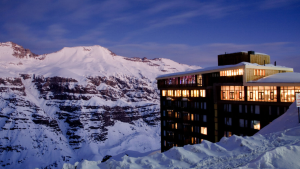 Hotel iluminado ao anoitecer em meio às montanhas nevadas dos Andes, com céu azul-escuro e neve acumulada ao redor.