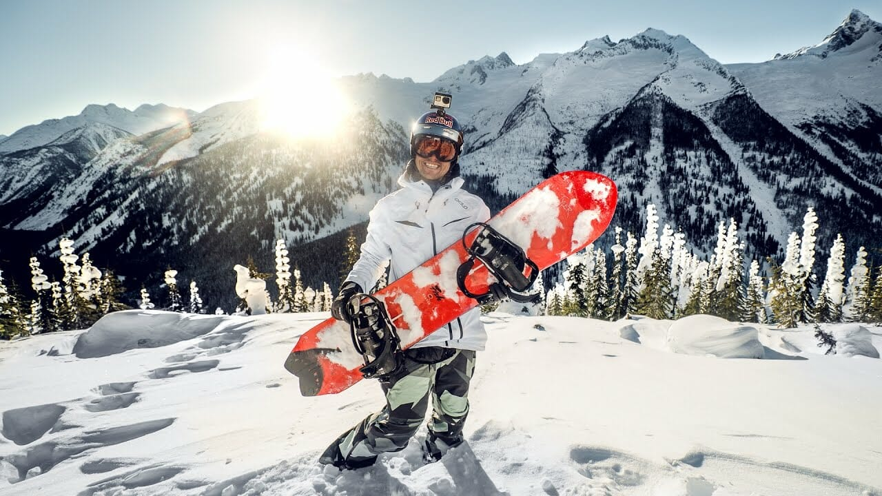 Pessoa segurando uma prancha de snowboard vermelha sobre a neve, com capacete e câmera GoPro, em meio a uma paisagem de montanhas nevadas ao pôr do sol.