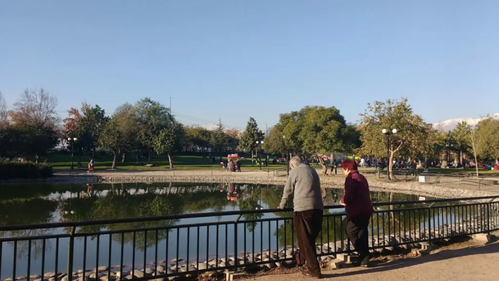 Duas pessoas apoiadas na grade observam o lago de um parque; ao fundo, famílias passeiam e há montanhas nevadas sob céu azul.