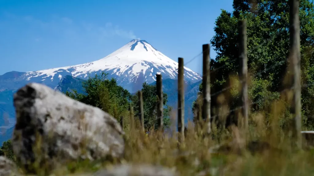 Vulcão coberto de neve visto ao fundo de uma cerca de madeira, em cenário natural do Chile.