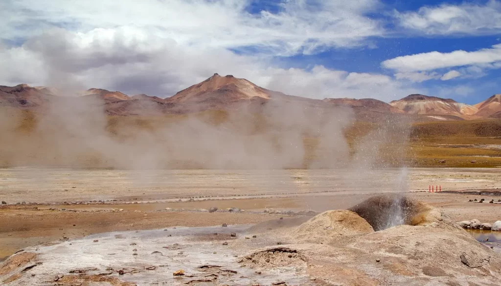 Gêiseres no Deserto do Atacama com montanhas ao fundo, liberando vapor sob o céu parcialmente nublado.