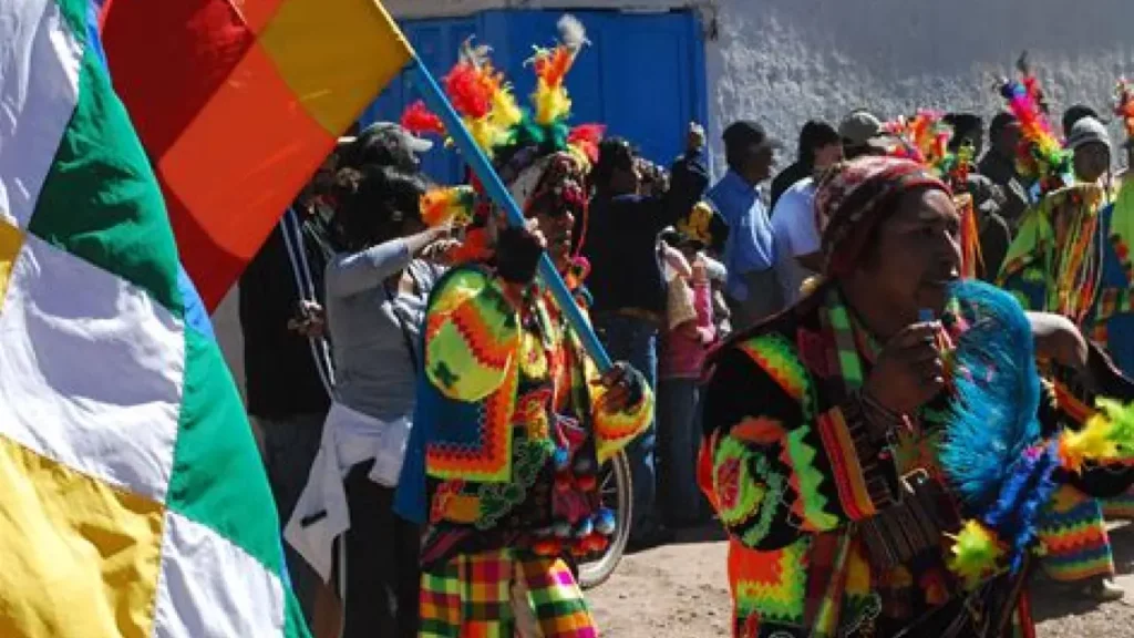 Uma imagem vibrante de uma celebração cultural andina, onde pessoas vestidas com trajes tradicionais coloridos participam de uma festa. A bandeira Wiphala, símbolo de várias comunidades indígenas dos Andes, está em destaque, representando a diversidade e a unidade cultural. Os trajes são ricos em cores e padrões, refletindo a herança cultural e a importância das tradições festivas na comunidade. A atmosfera é alegre, com música e danças típicas que fazem parte integral das festividades.