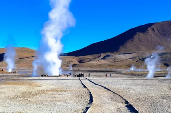 Gêiseres de vapor branco emergindo do solo na paisagem árida dos Gêiseres del Tatio no Deserto do Atacama, com montanhas ao fundo sob um céu azul claro. Turistas caminham ao longo de trilhas de terra para observar de perto o fenômeno natural.