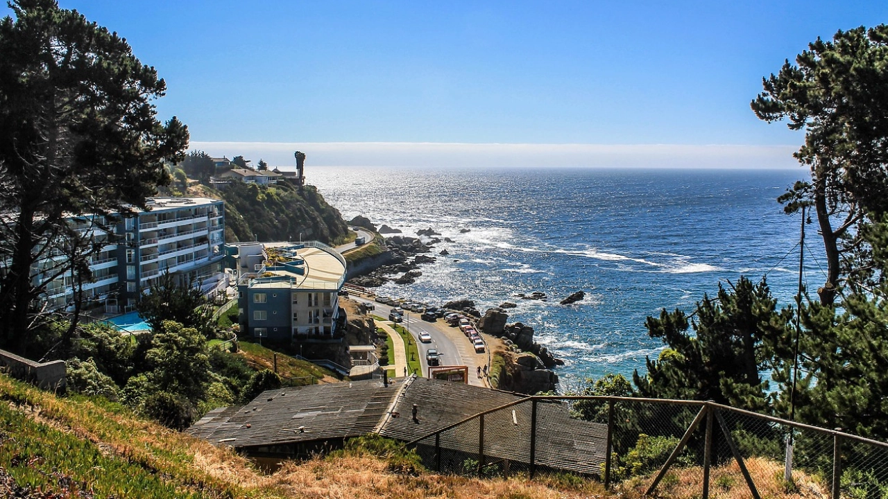 Vista da costa de Viña del Mar, Chile, com o oceano Pacífico ao fundo, uma estrada costeira sinuosa e edifícios modernos à beira do penhasco. O cenário é cercado por vegetação e o mar azul profundo, refletindo a luz do sol.