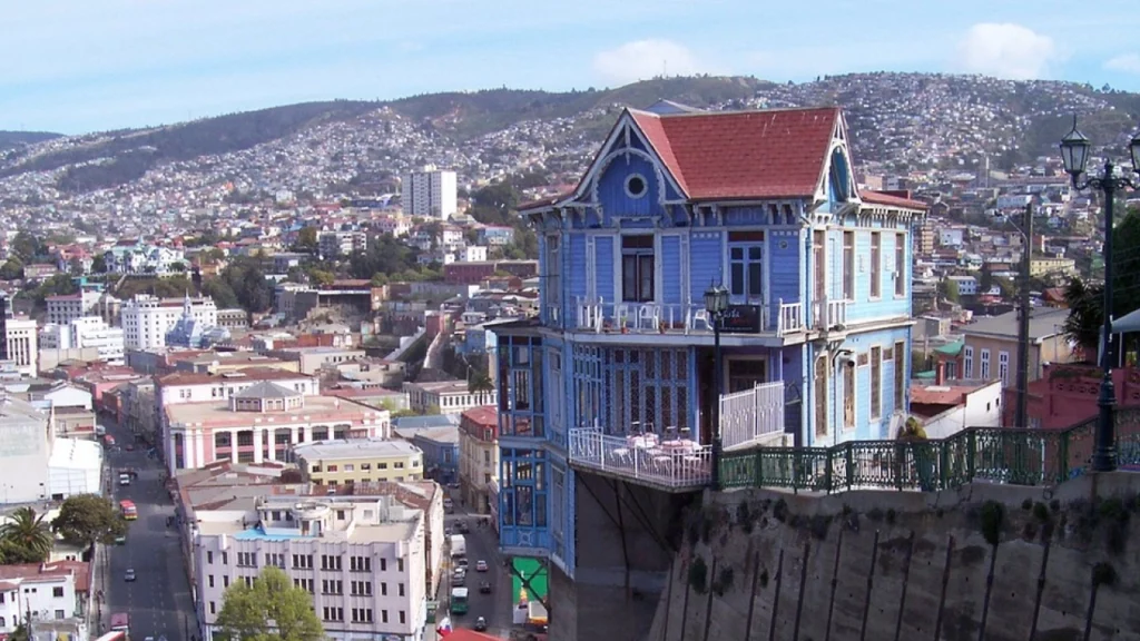 Vista de uma casa colorida em Valparaíso, Chile, situada em uma encosta com um funicular vermelho ao lado. A cidade ao fundo é repleta de construções nas colinas, um exemplo típico da arquitetura vibrante e das paisagens únicas de Valparaíso.