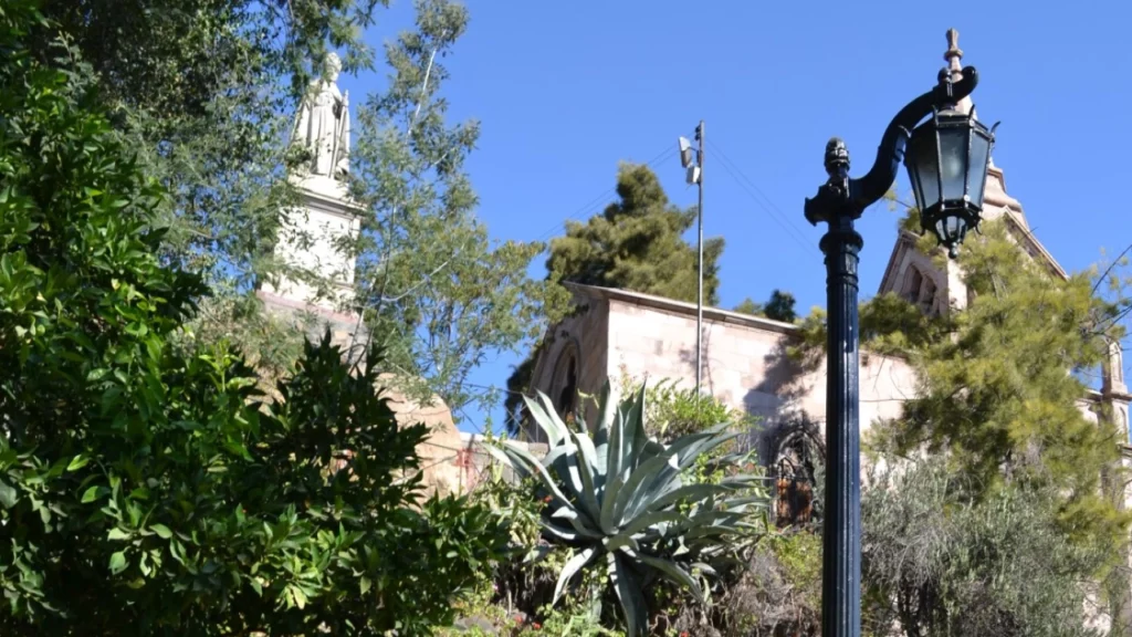 Um parque verde exuberante com uma variedade de plantas e árvores, emoldurado por uma construção histórica e um poste de luz ao centro.