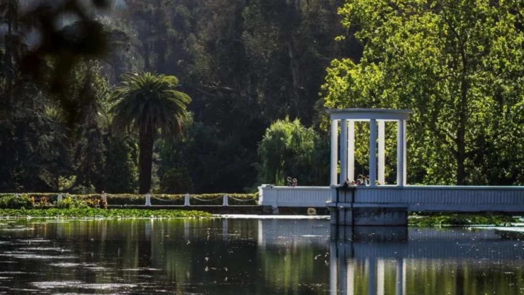 Um lago sereno refletindo o verde das árvores ao redor, com uma estrutura branca em forma de colunas ao fundo, criando uma paisagem pacífica.