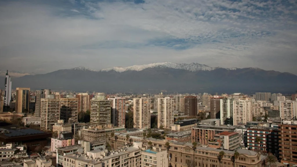 Vista panorâmica de Santiago, Chile, com prédios modernos ao fundo e a Cordilheira dos Andes coberta de neve no horizonte.