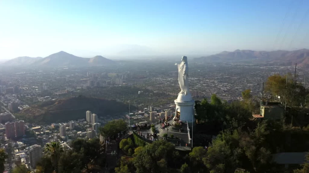 Vista panorâmica do Cerro San Cristóbal em Santiago, Chile, com a estátua da Virgem Maria ao fundo e a cidade se estendendo no horizonte.