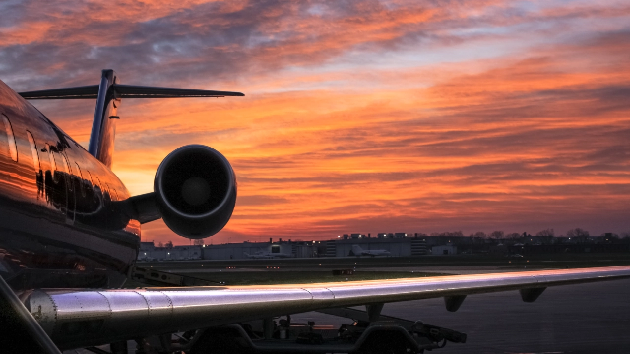 Avião em um aeroporto durante o pôr do sol, com o céu alaranjado refletido na fuselagem.