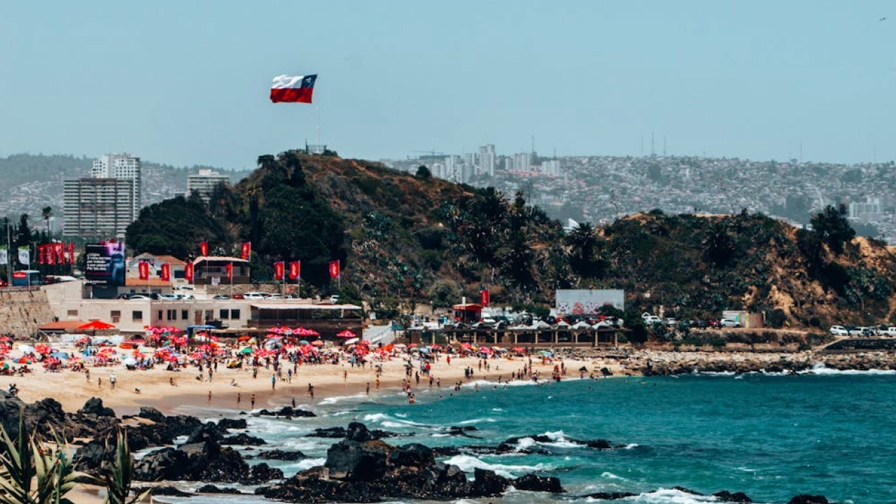 Praia movimentada em Viña del Mar, Chile, com bandeira chilena ao fundo, pessoas aproveitando o sol e o mar azul.