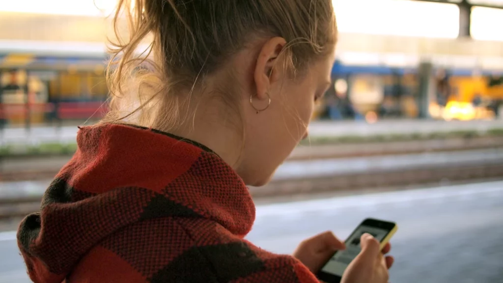 Mulher usando um moletom xadrez vermelho, olhando para a tela do celular enquanto espera na plataforma de uma estação de trem.