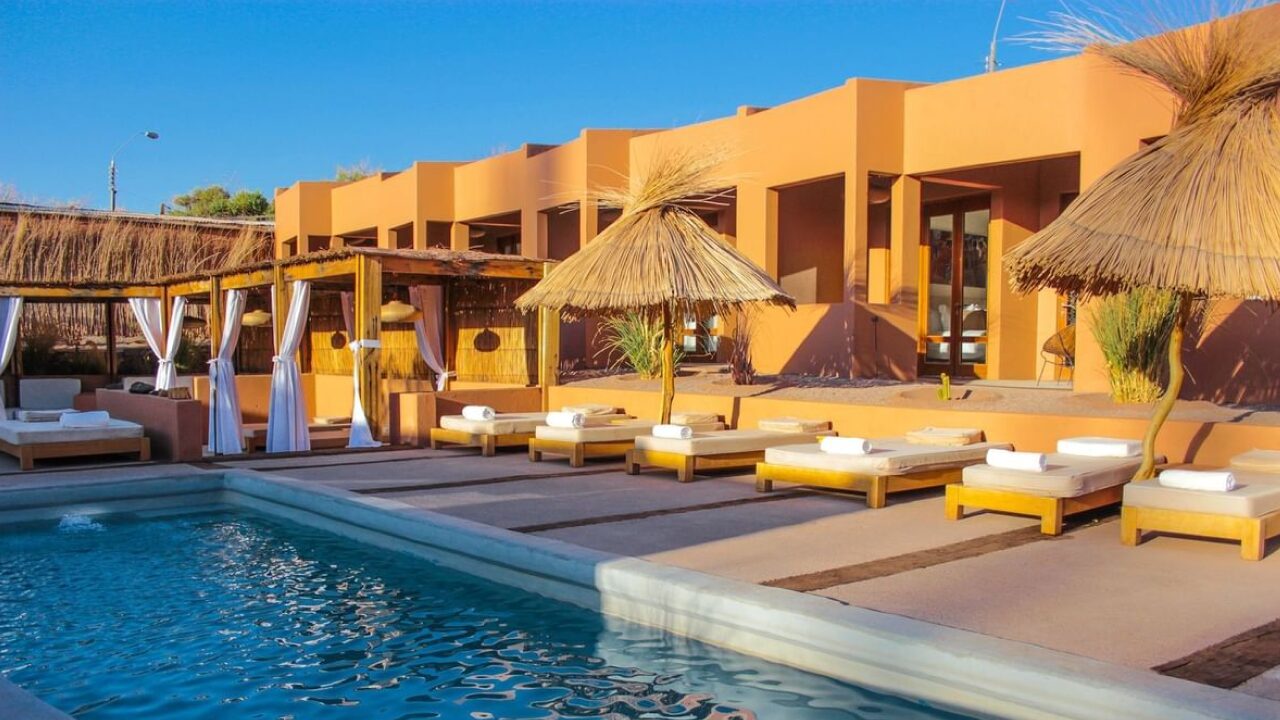 Área externa de um hotel em San Pedro de Atacama, com piscina ao ar livre rodeada por espreguiçadeiras e cabanas com cobertura de palha, em um cenário desértico e ensolarado.