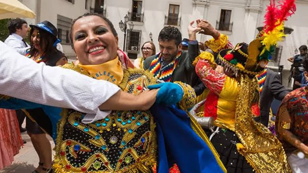 Dançarinos em trajes tradicionais coloridos celebram em uma praça de Santiago, Chile, durante uma festa cultural, com música, alegria e expressões vibrantes.