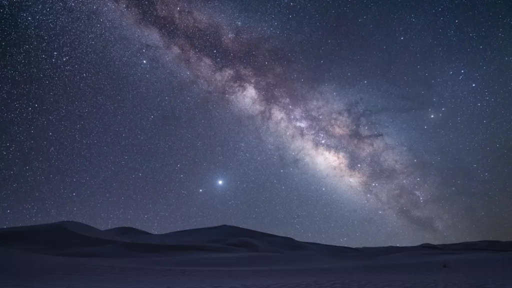 Céu estrelado no Deserto do Atacama com a Via Láctea brilhando intensamente acima das dunas, em uma visão surreal e inesquecível.