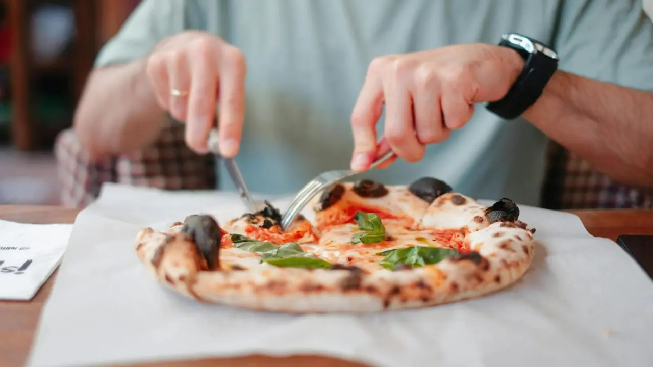 Pessoa cortando uma pizza gourmet com tomate e manjericão fresco, utilizando faca e garfo, em uma mesa de madeira.