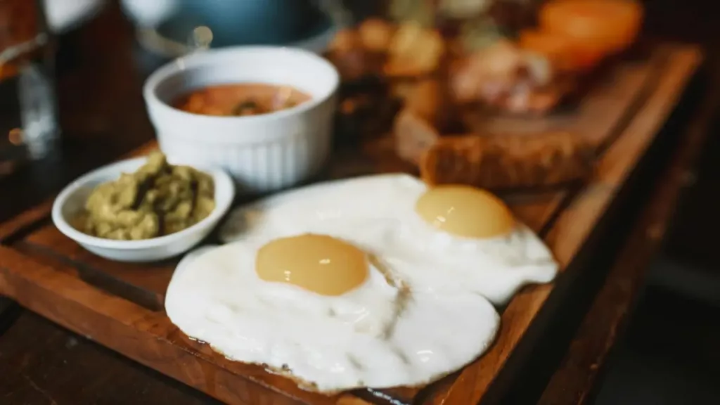 Prato de café da manhã com ovos fritos e acompanhamentos como guacamole e molho, servido em tábua de madeira.