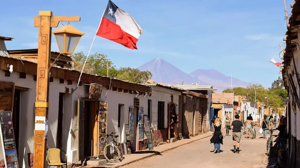 Rua principal de San Pedro de Atacama, com bandeiras do Chile, casas de adobe e o vulcão Licancabur ao fundo em um dia ensolarado.
