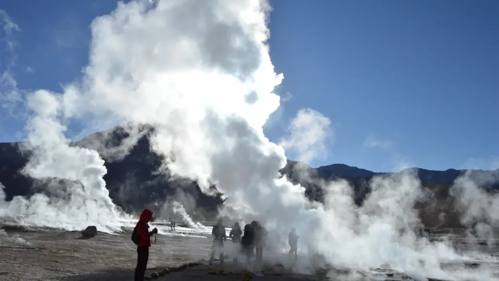 Turistas observando a atividade dos Geysers del Tatio no deserto do Atacama, cercados por colunas de vapor sob céu azul e montanhas ao fundo.