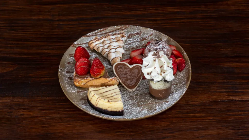 Prato de sobremesas com morangos frescos, pedaços de cheesecake, waffle coberto com açúcar de confeiteiro, crepe, copinho de chocolate com chantilly e calda de chocolate em formato de coração, servido sobre prato dourado com decoração de açúcar, em uma mesa de madeira escura.