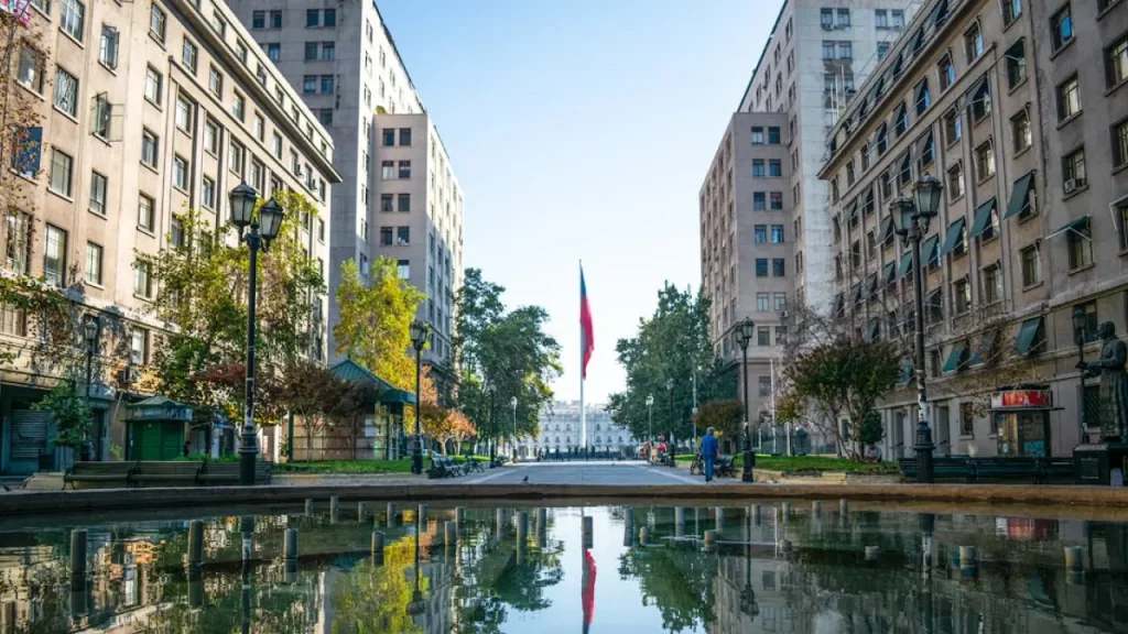 Centro histórico de Santiago com bandeira chilena ao fundo refletida em espelho d’água, entre prédios clássicos.