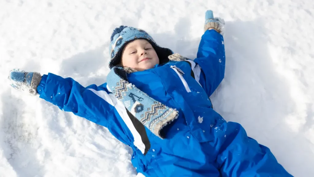 Criança deitada na neve vestindo roupa de inverno azul, gorro e cachecol combinando, fazendo anjo de neve com os braços abertos.