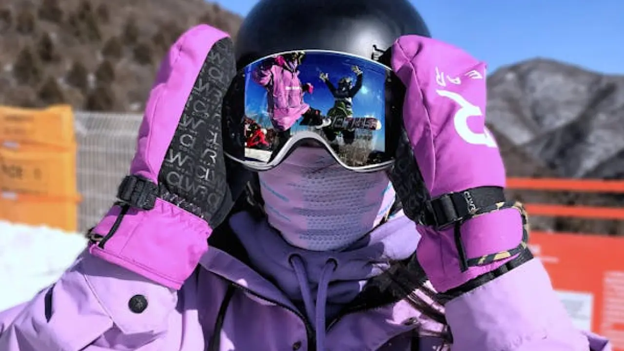 Persona con ropa de esquí color morado y guantes rosas, ajustando sus gafas reflejantes donde se ve a otros esquiadores en acción.