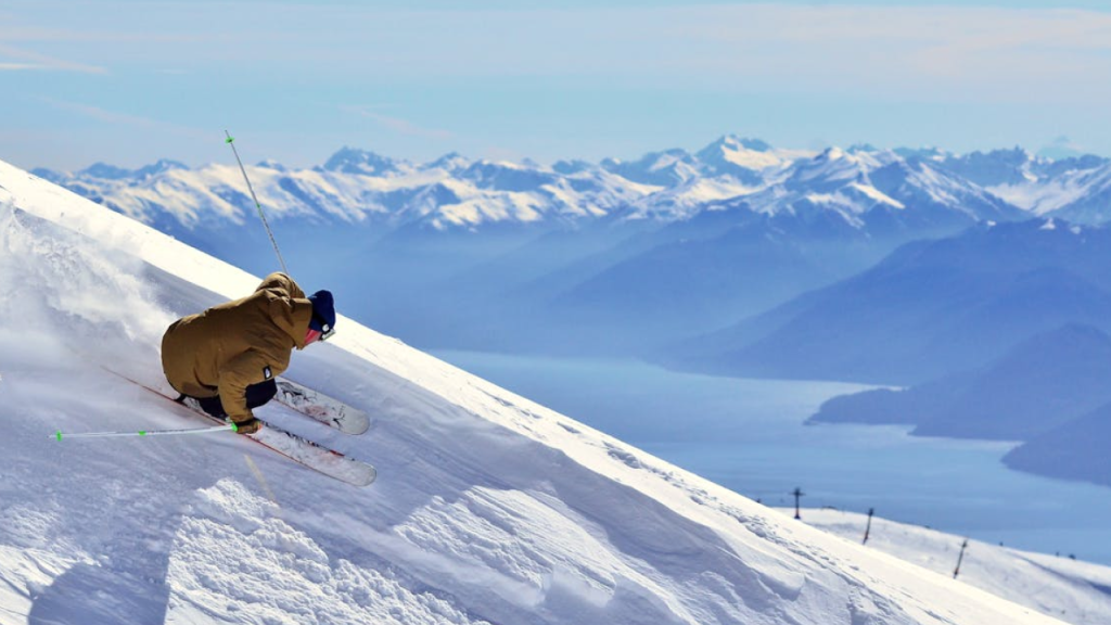 Esquiador descendo encosta íngreme com cordilheira e lago ao fundo, mostrando a emoção de conhecer a neve no Chile.