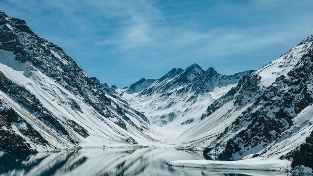 Vista panorâmica das montanhas nevadas da Cordilheira dos Andes no inverno chileno, com lago espelhado em destaque.
