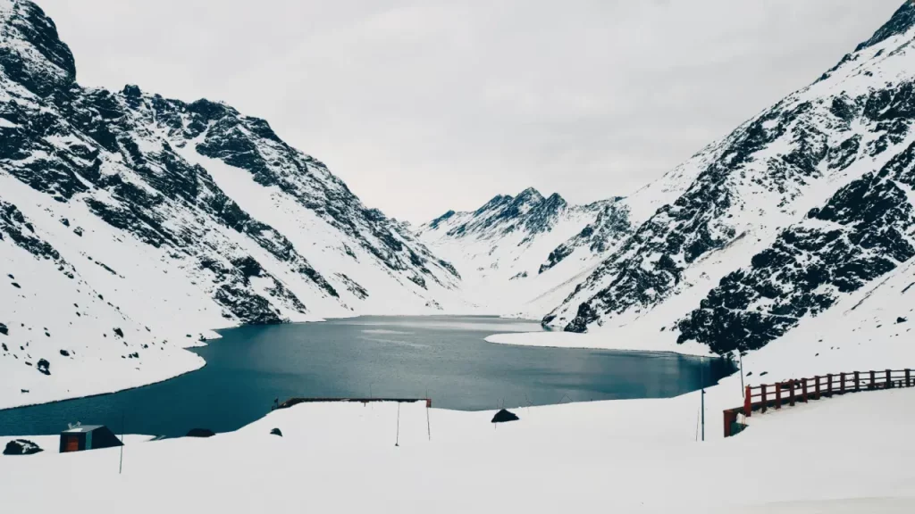 Lago cercado por montanhas cobertas de neve, com águas calmas refletindo o céu nublado, em uma paisagem andina de inverno.
