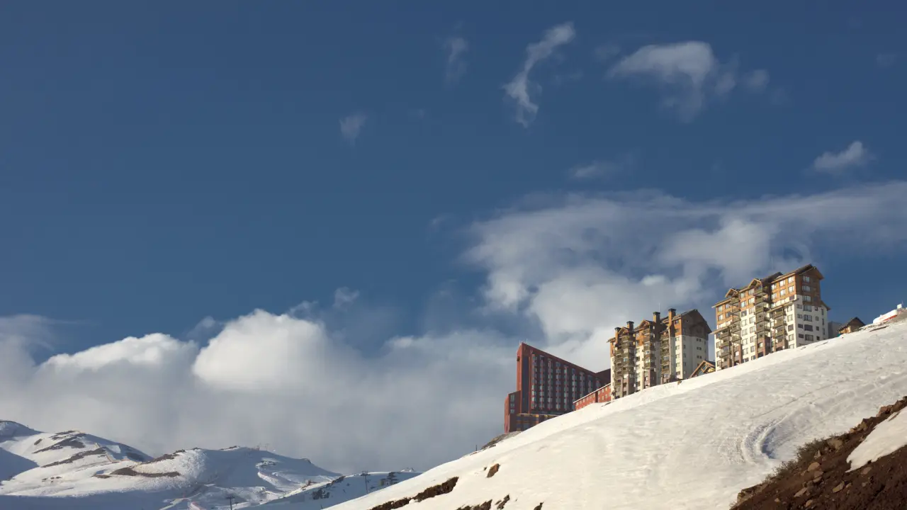 Edificios de hoteles en lo alto de una pendiente nevada, bajo un cielo azul despejado en una estación de esquí andina.