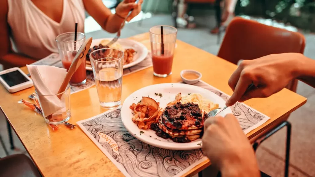 Duas pessoas tomando café da manhã em restaurante, com panquecas, ovos e suco na mesa.