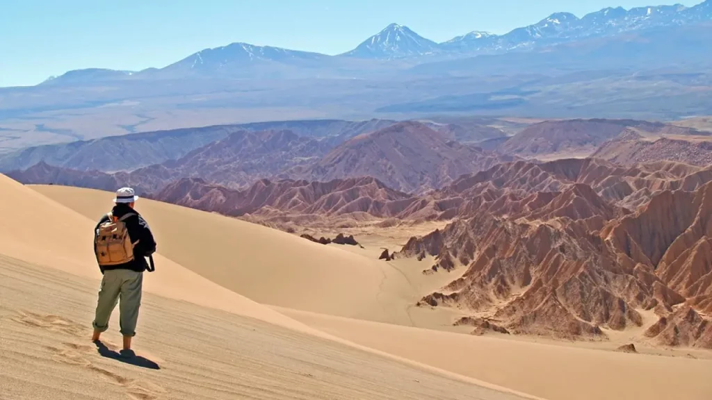 Viajante com mochila observa as montanhas e dunas do Deserto do Atacama em um dia claro, com vulcões ao fundo.