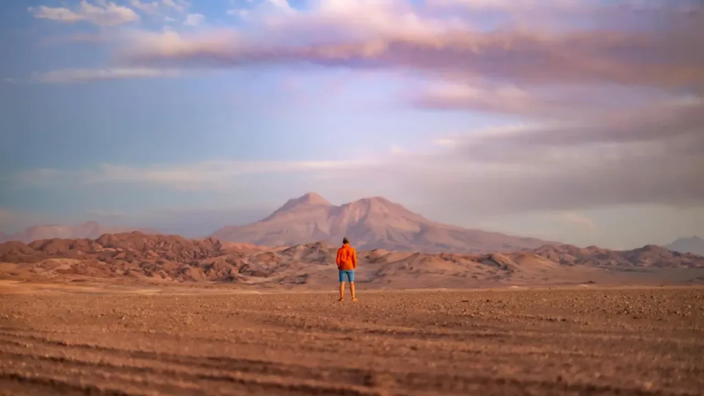 Um viajante de costas, usando um casaco laranja, admirando a vastidão do deserto do Atacama com montanhas ao fundo, ao entardecer.