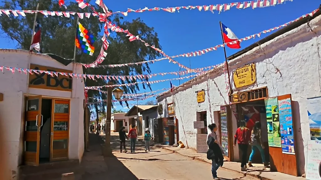 Rua principal de San Pedro de Atacama decorada com bandeirinhas, bandeiras do Chile e da Wiphala, com turistas caminhando em frente a lojas e agências de turismo locais.