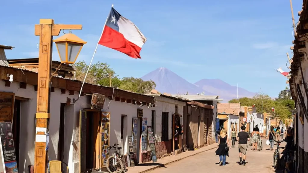 Vista da rua principal de San Pedro de Atacama com bandeiras do Chile, turistas caminhando e o vulcão Licancabur ao fundo em dia ensolarado.