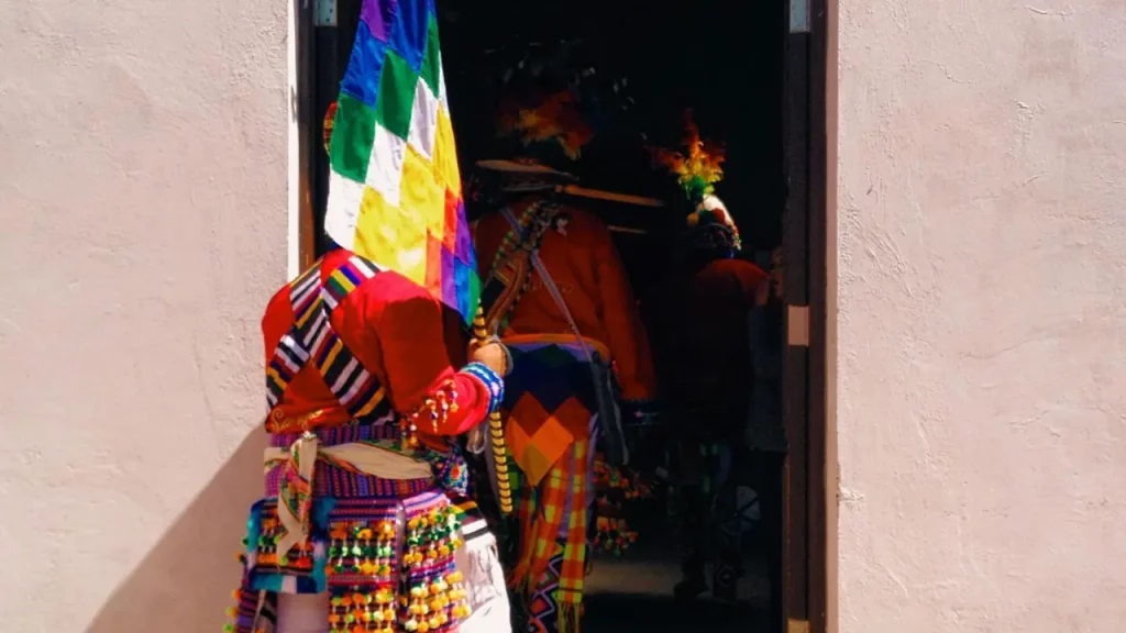 Pessoa em trajes típicos andinos entra em uma construção durante uma celebração cultural em San Pedro de Atacama, carregando a bandeira Wiphala.