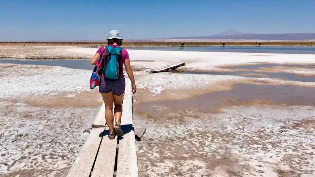 Uma pessoa caminhando sobre uma passarela de madeira em meio ao deserto de sal no Atacama, com um cenário árido e montanhas ao fundo.