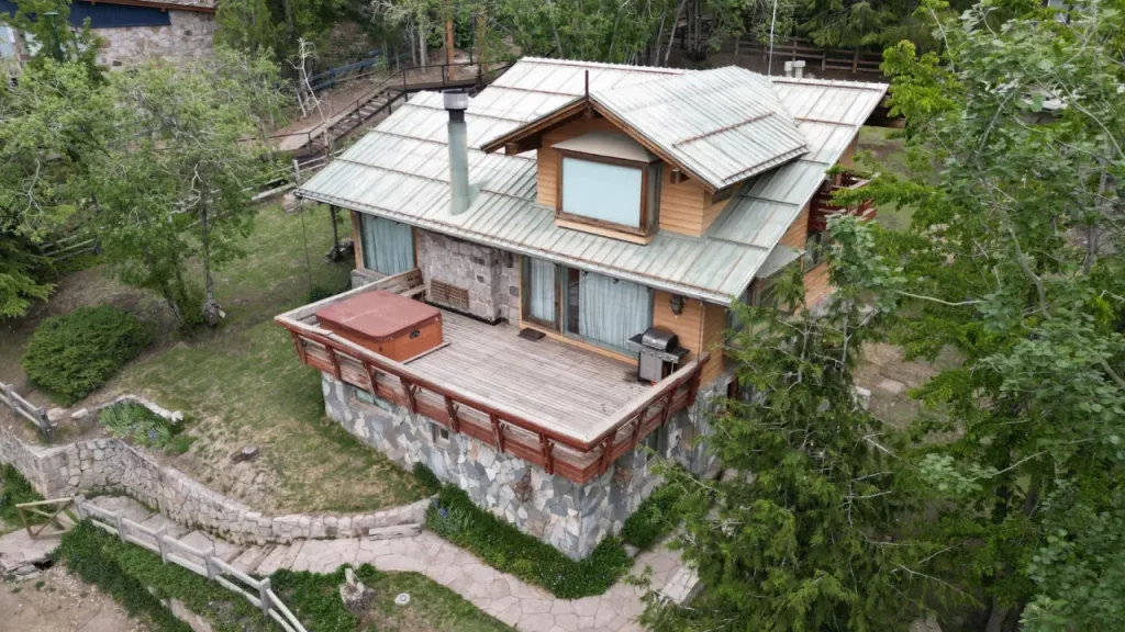 Fachada da Casa Condor, hostel nas montanhas do Chile, com deck de madeira, jacuzzi externa e rodeada por árvores verdes.